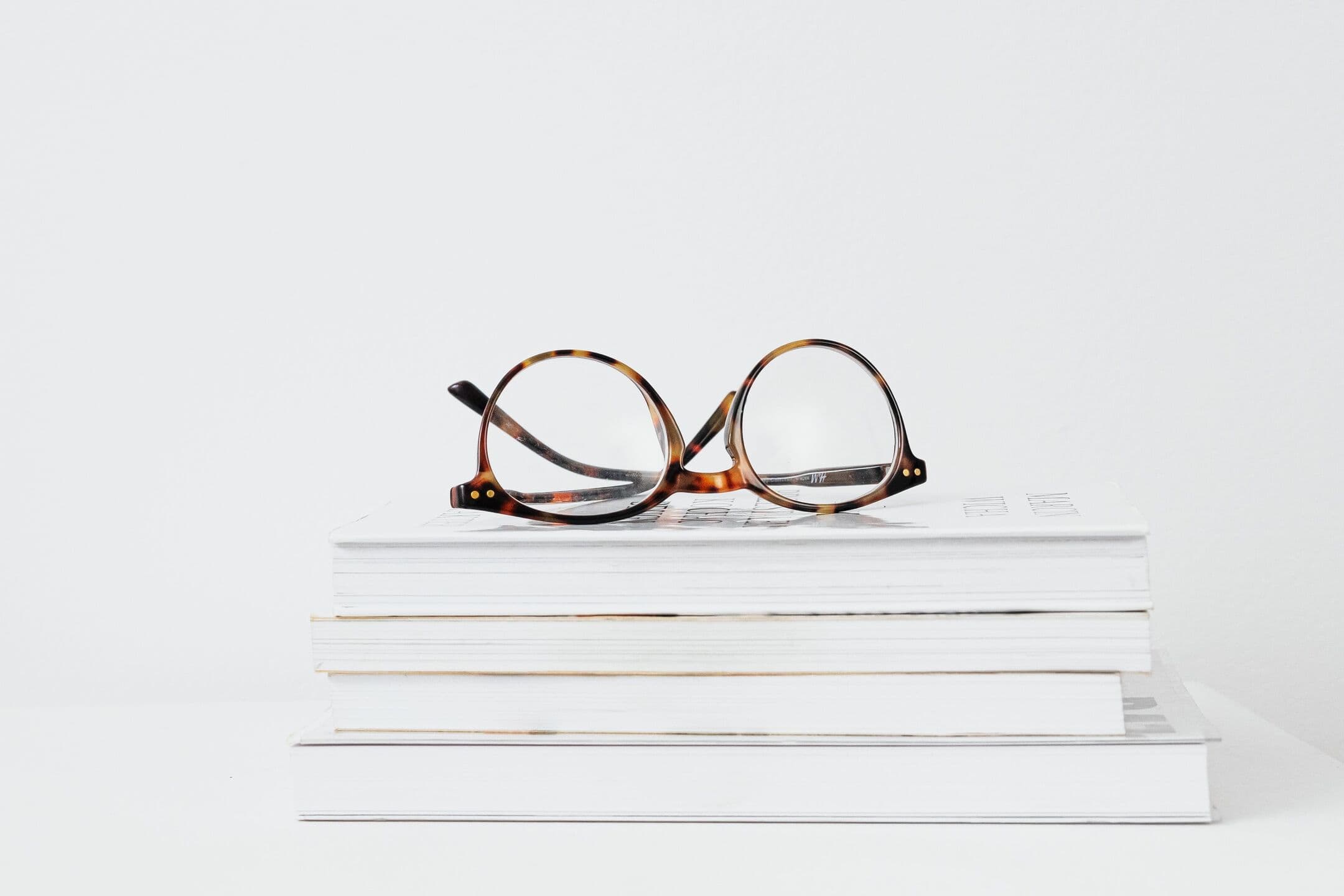 Tortoiseshell reading glasses resting on a small stack of white books against a white wall