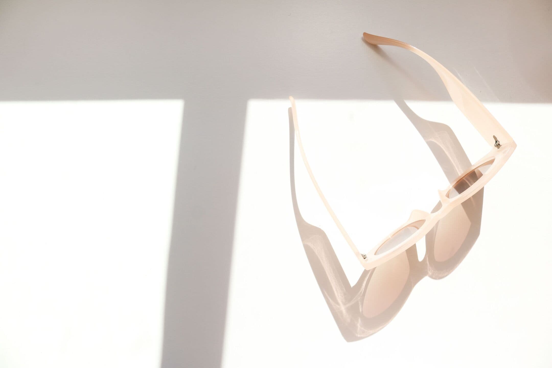 Pale sunglasses resting on a white surface in bright window light, casting a soft shadow