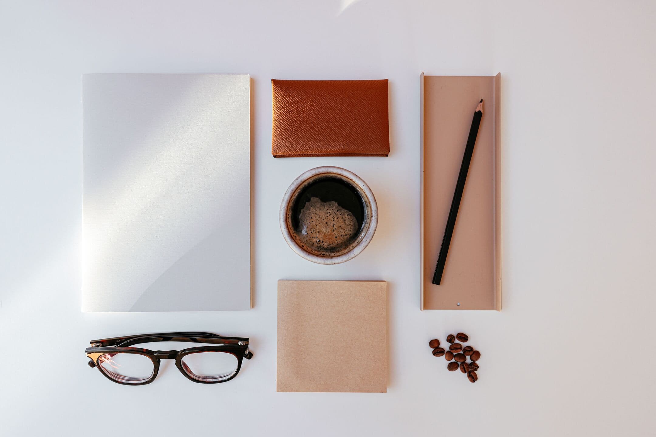 A blank folder, a leather card wallet, a cup of coffee, reading glasses, and a pencil laid out on a white desk in window light