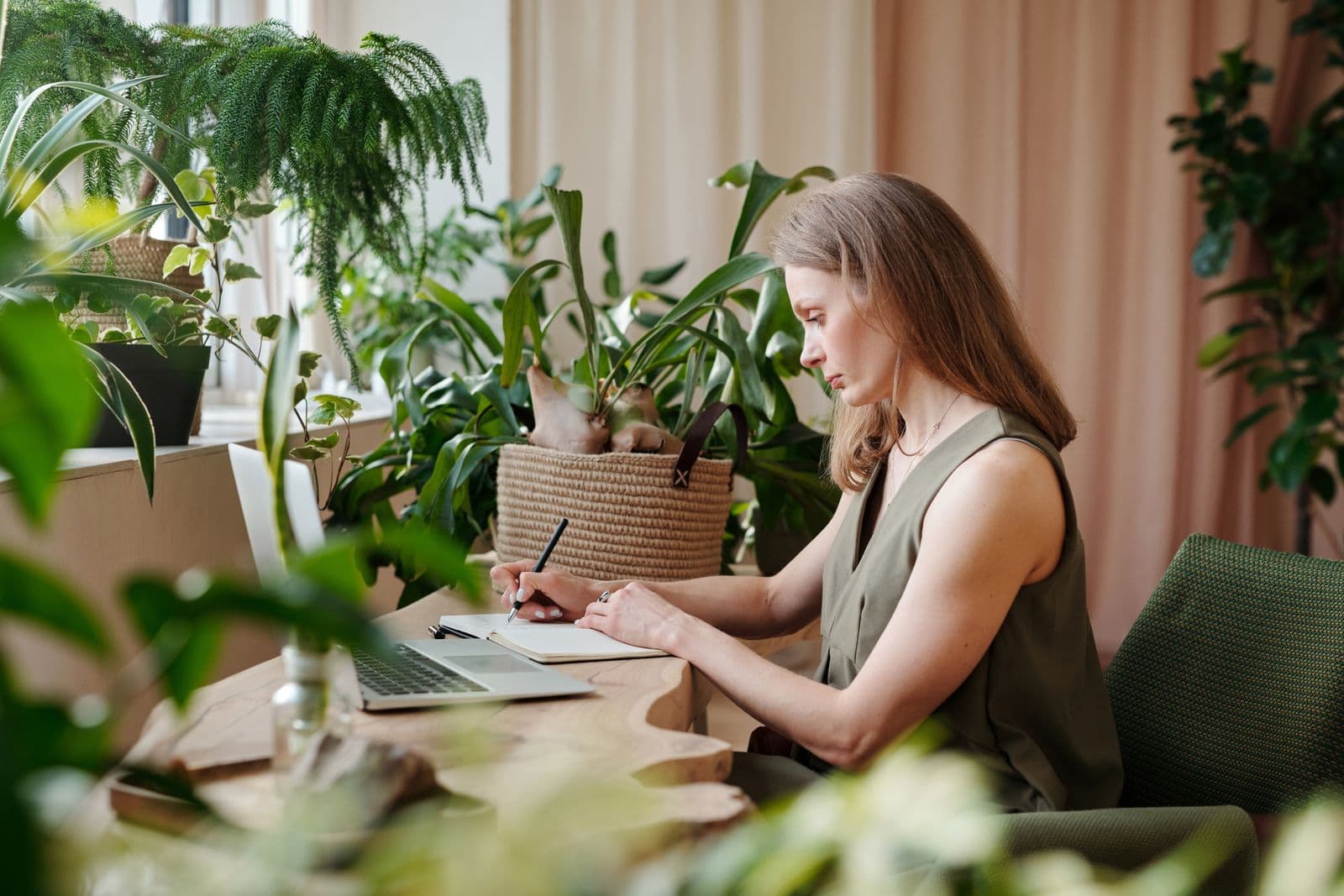 Woman writing in a notebook at a desk surrounded by houseplants