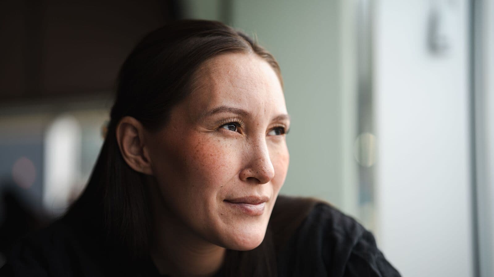 Dark-haired woman in a black sweater gazing out of frame with a slight smile in soft window light