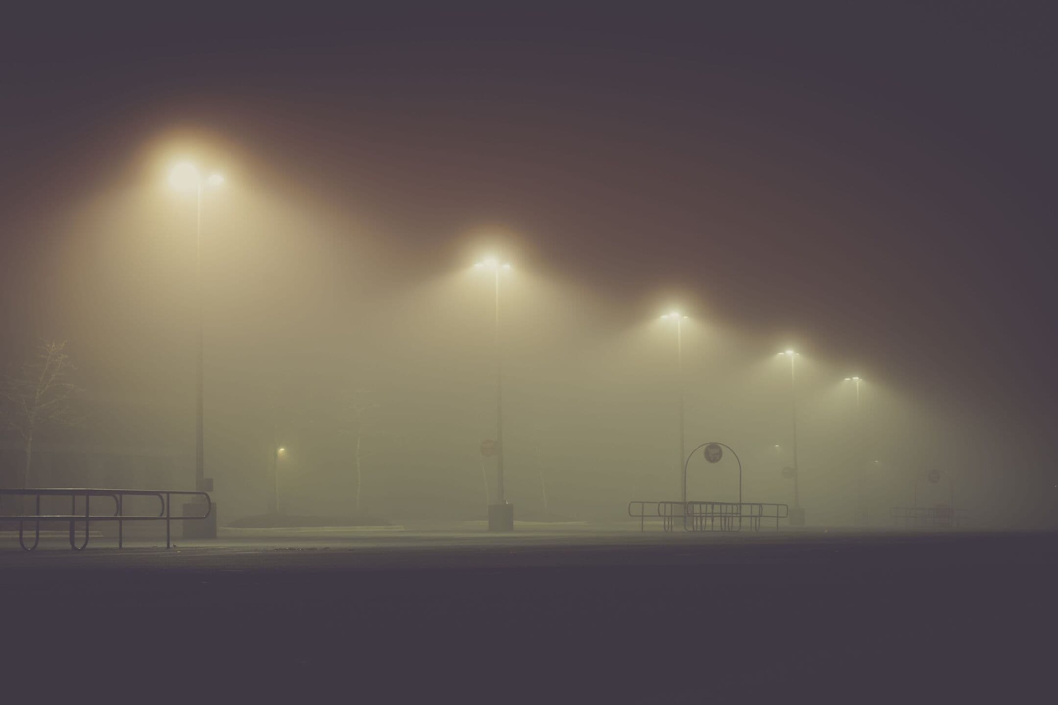 Lights on tall poles glowing with wide halos over an empty lot on a foggy night