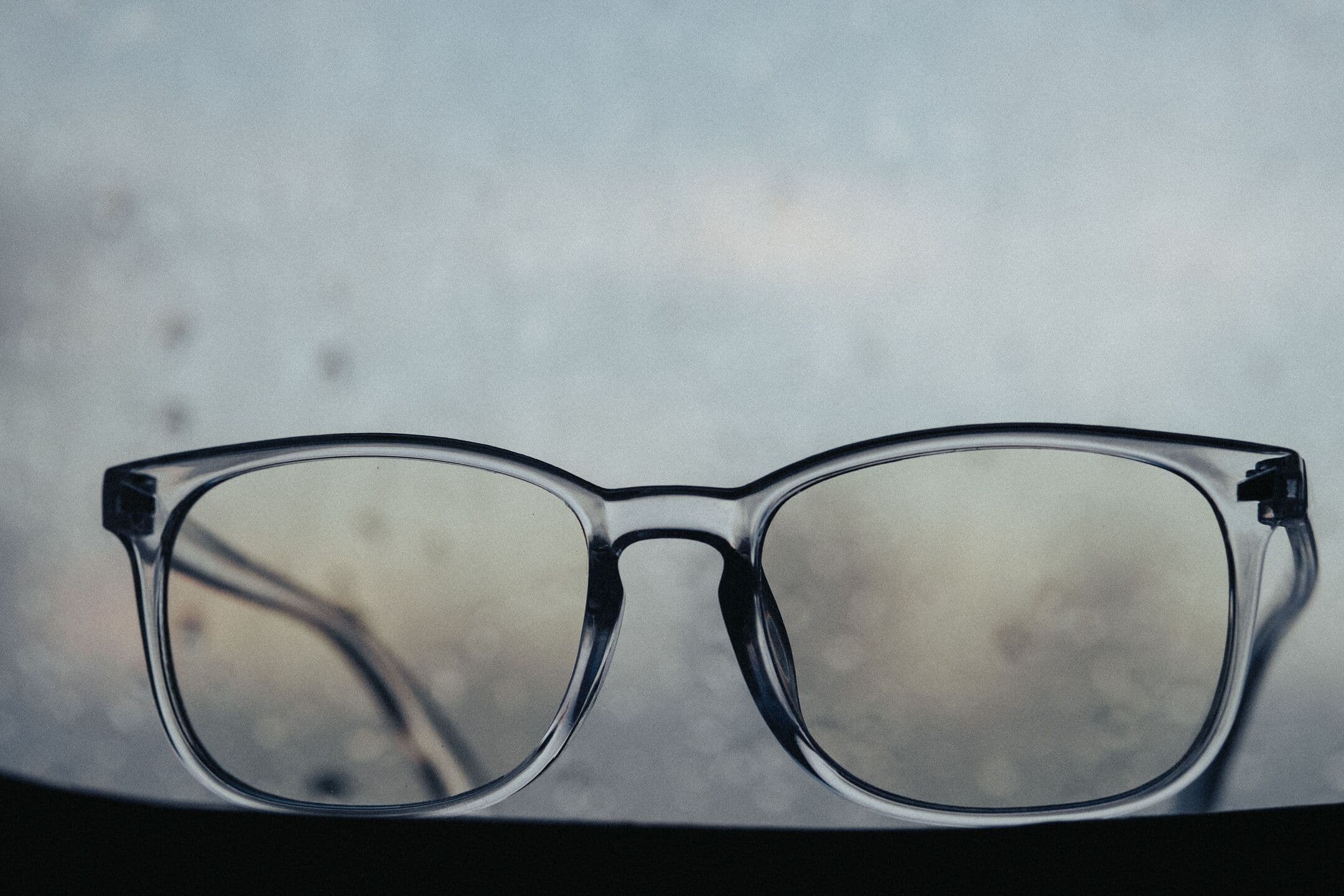 A pair of clear-framed glasses resting on a sill in front of a rain-streaked window