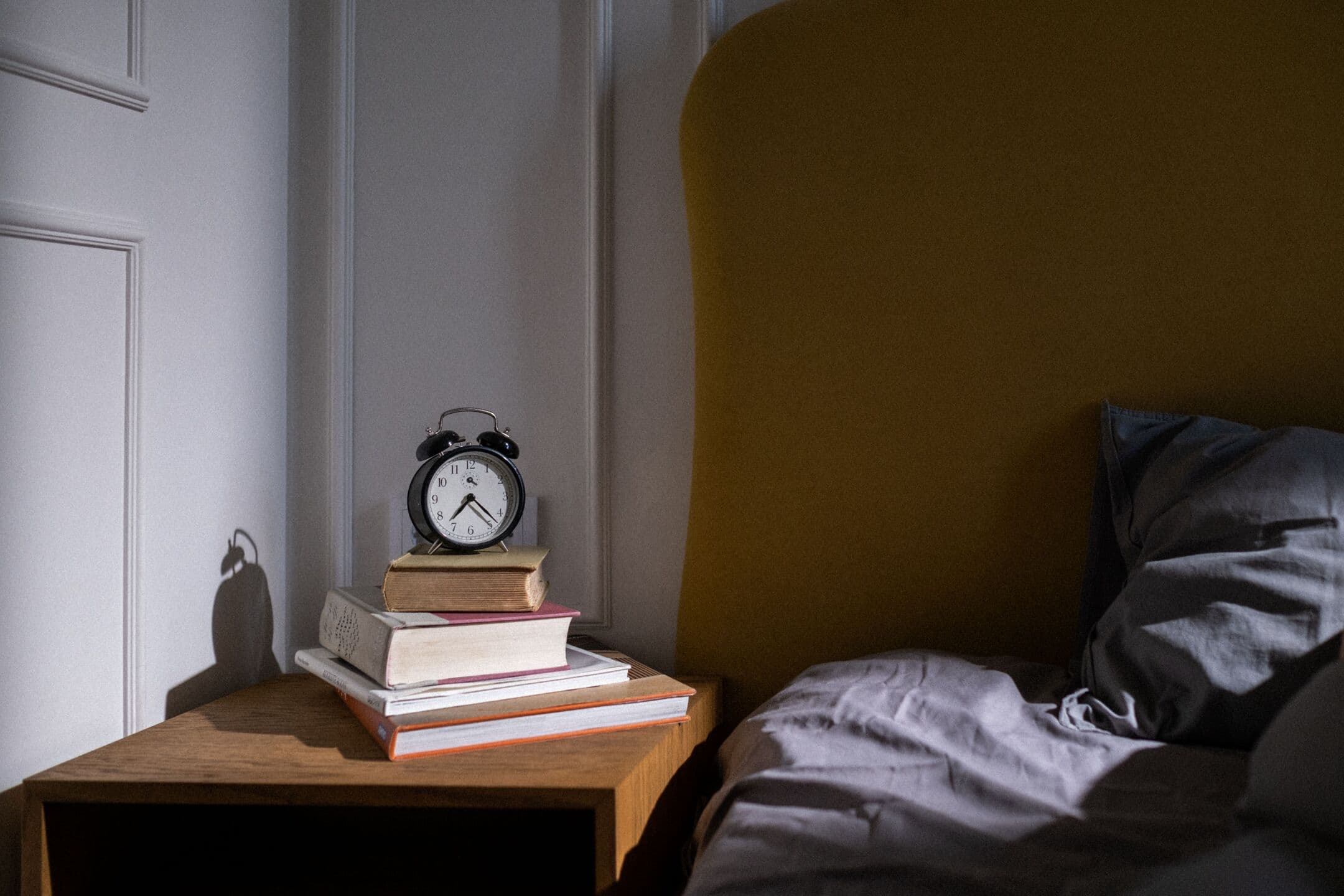 A twin-bell alarm clock on a stack of old books on a wooden nightstand beside an unmade bed