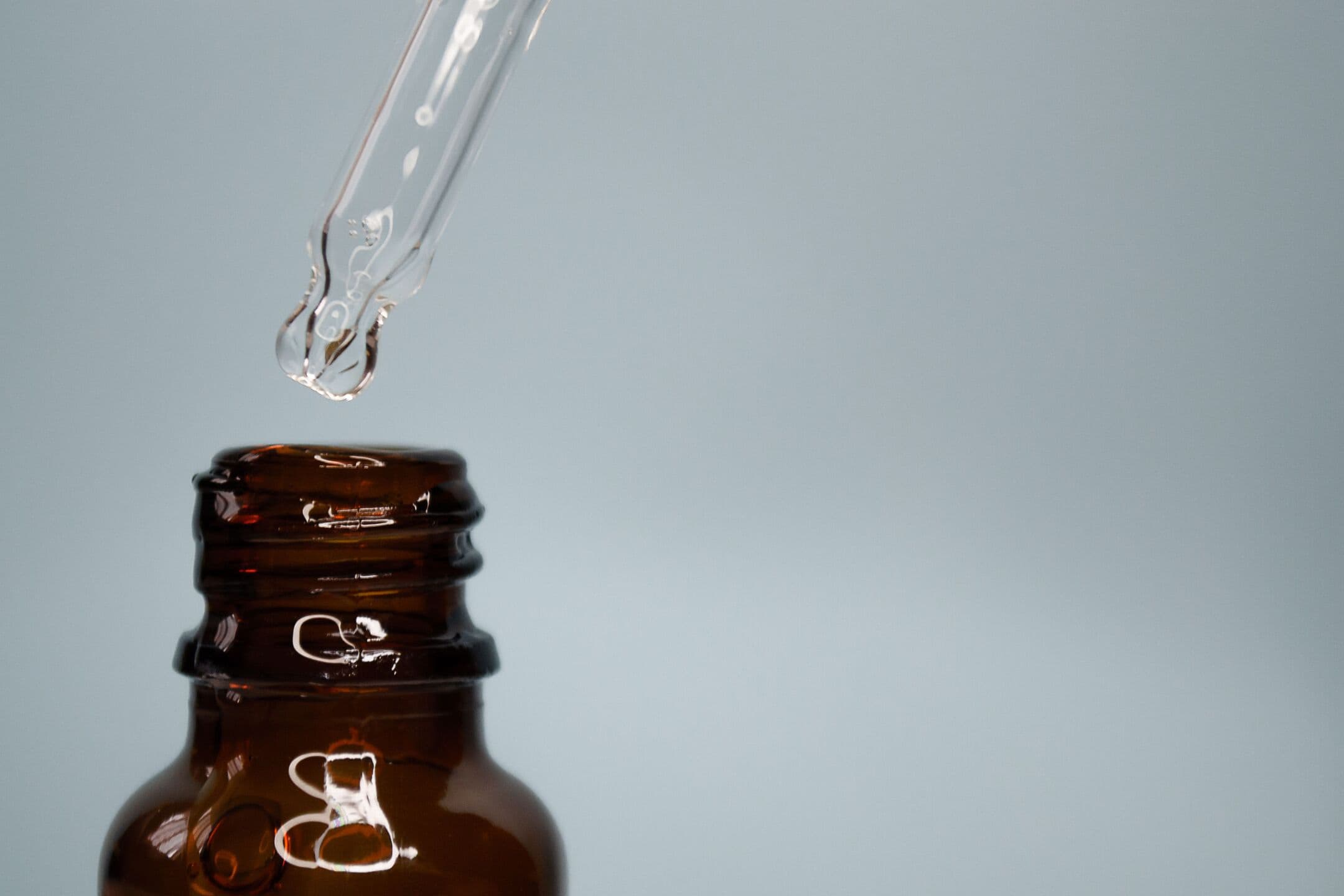 A glass dropper holding a single drop above an amber bottle, against a soft grey background