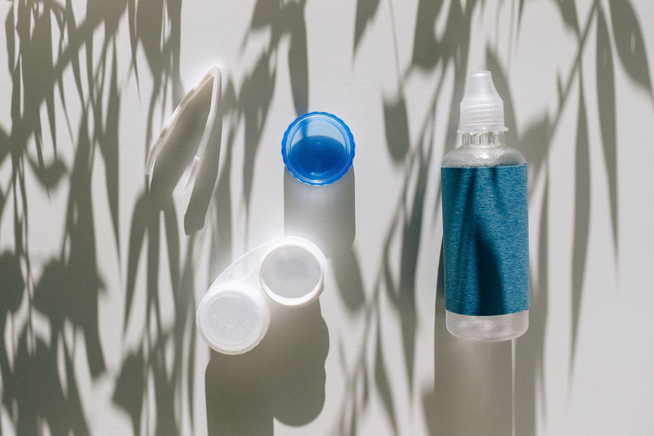 A contact lens case, its blue cap, and a bottle of solution on a white surface crossed by leaf shadows